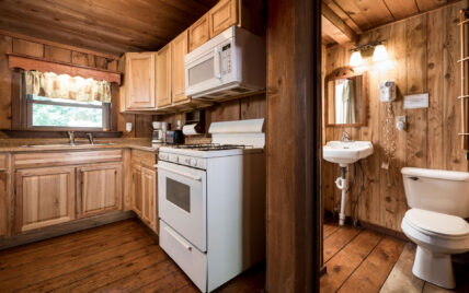 Kitchen and dining area with bathroom in the Liggett Cabin at Gateway Lodge in Cook Forest, Pennsylvania, featuring light wood cabinets, white appliances (refrigerator, microwave, stove), a round wooden dining table with four chairs, and wood-paneled walls and ceiling.
